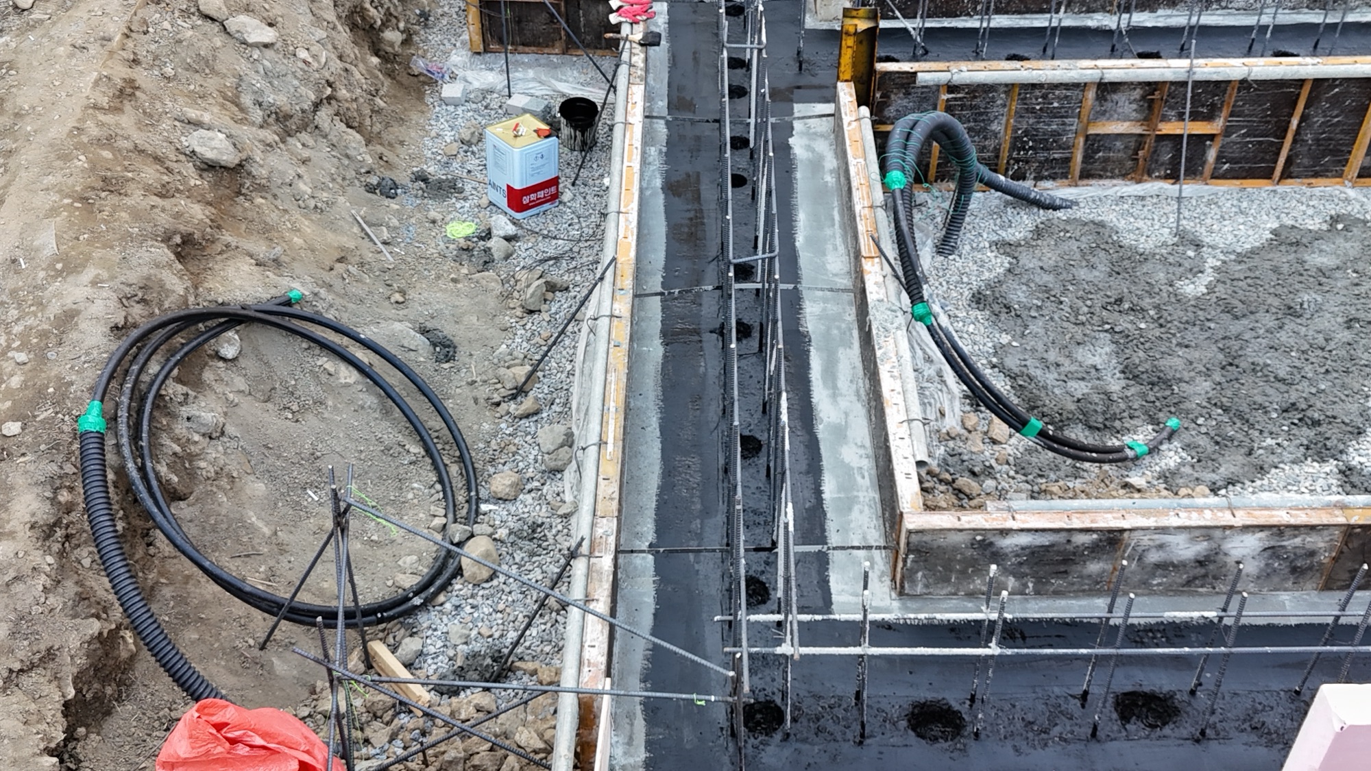 줄기초 푸터 시공 중의 기초 구조물 이미지, 철근과 배수관이 설치된 상태이며, 주변은 흙과 파쇄석으로 정리되어 있음.