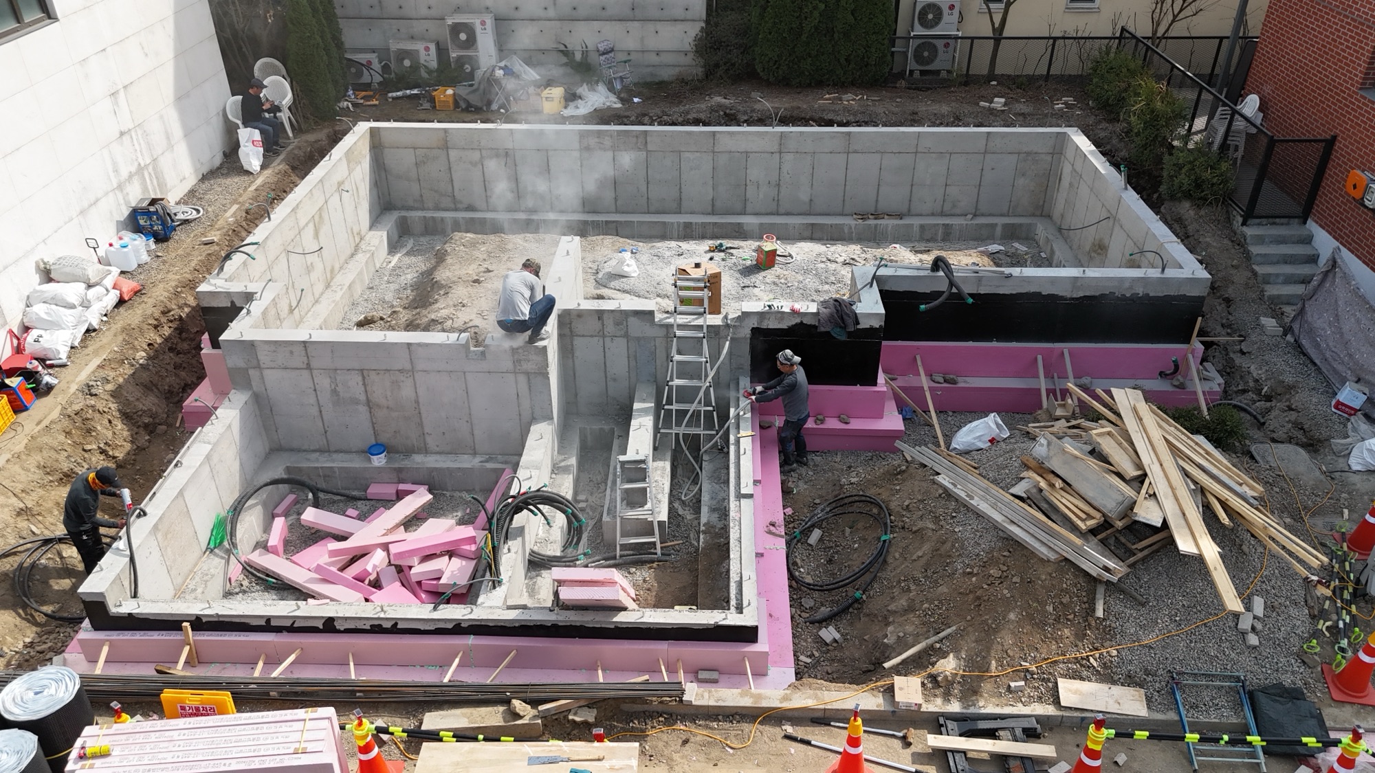 기초 공사가 진행 중인 건축 현장으로, 줄기초 구조물이 세워지고 있으며 고립된 부분에 방수 및 단열 재료가 사용되고 있다. 작업자들이 시공 중인 모습과 주변 자재들이 보인다.