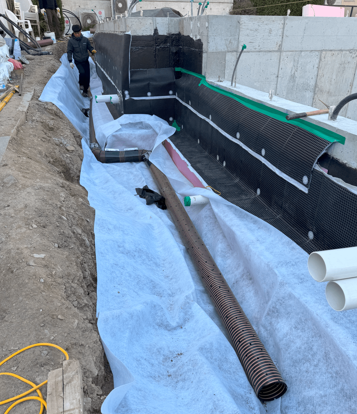 기초 공사 현장에서 프렌치 드레인 설치를 위한 준비 작업을 진행 중인 모습. 바닥에는 드레인 및 방수 재료가 깔려 있으며, 작업자가 서서 작업하고 있다.