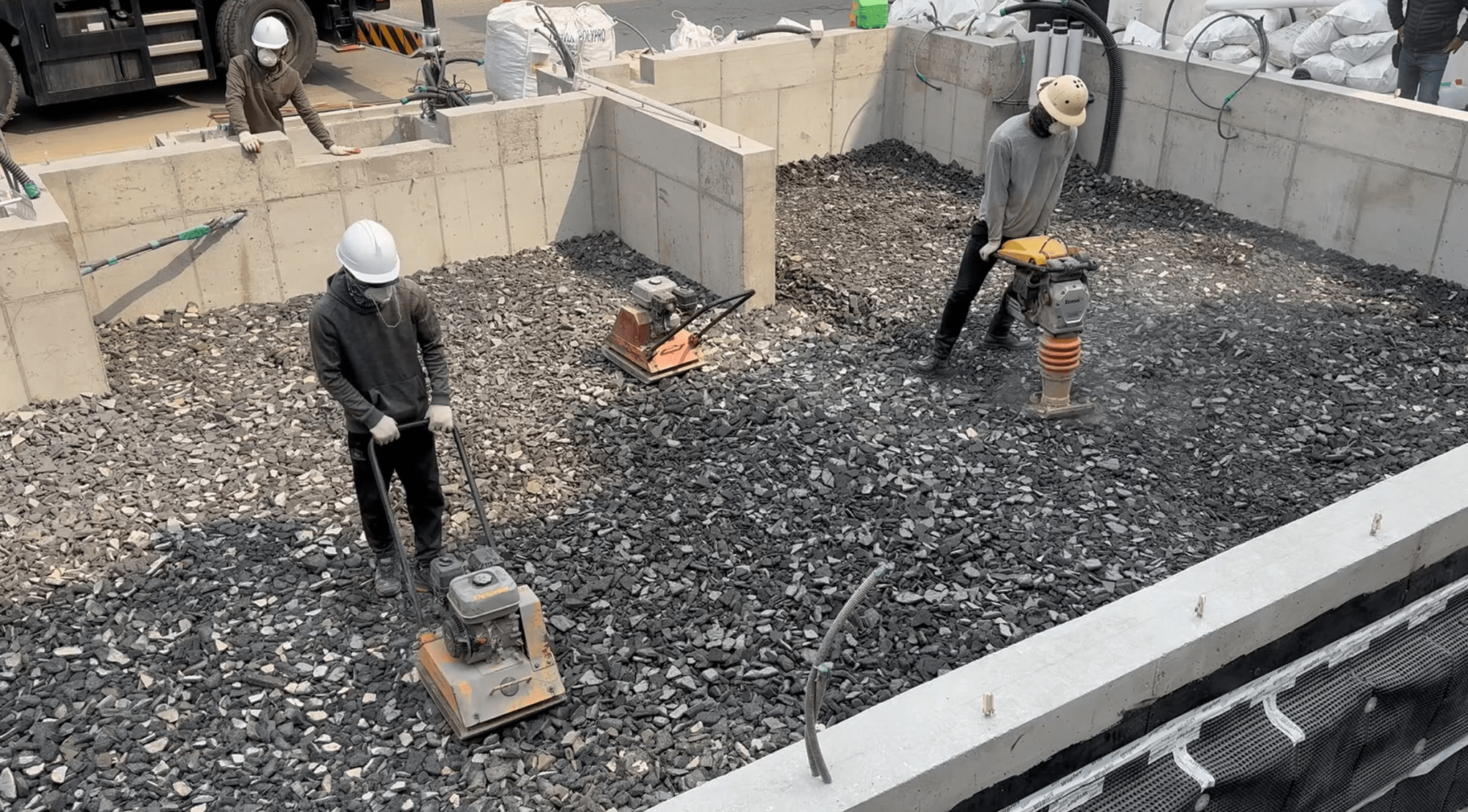 Construction workers using compactors on a gravel-filled foundation site, surrounded by concrete walls.