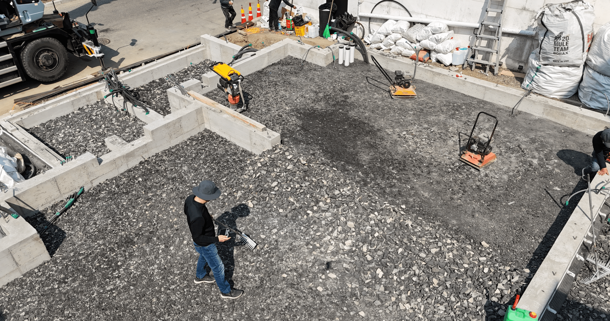 A construction site showing a foundation being prepared with crushed stone. Workers are using compactors, and various equipment is visible on the site.
