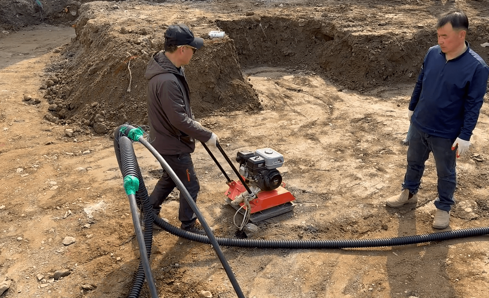 건설 현장에서 콤팩터를 사용하여 땅을 다지고 있는 작업자가 있는 장면.