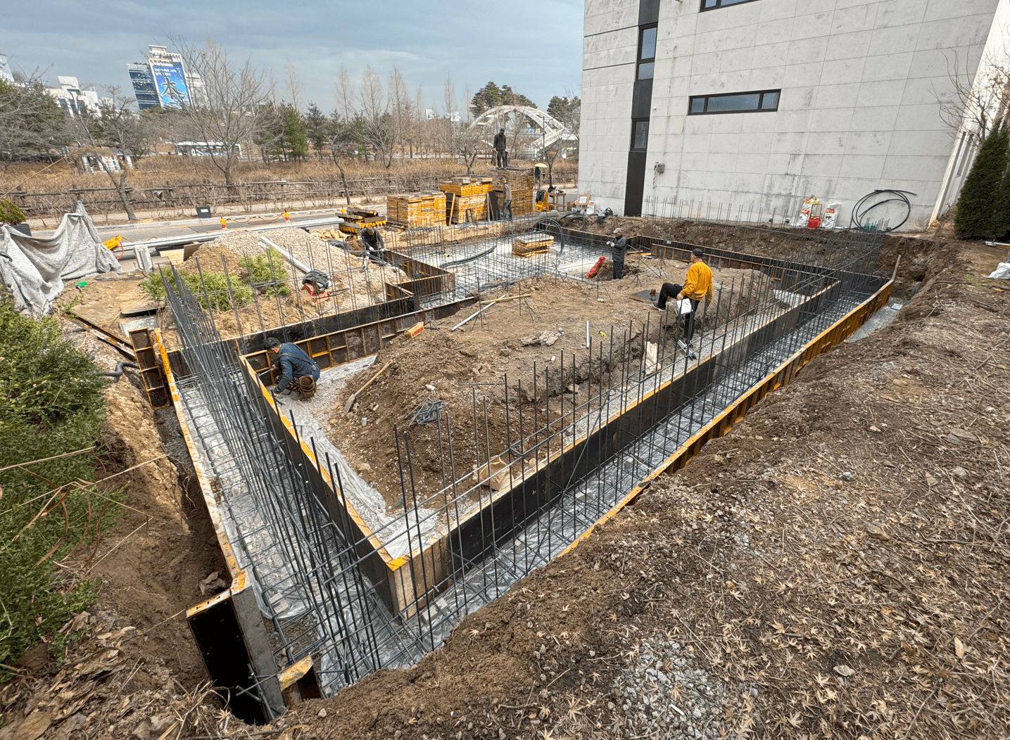건축 현장에서 기초 공사를 진행 중인 모습. 여러 작업자가 줄기초를 위한 거푸집에 철근을 배치하고 있으며, 주변에는 흙과 자재들이 보입니다.