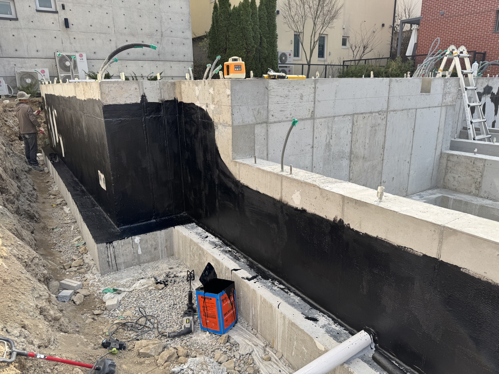 Concrete foundation wall with waterproofing applied, showing excavation and construction tools nearby.