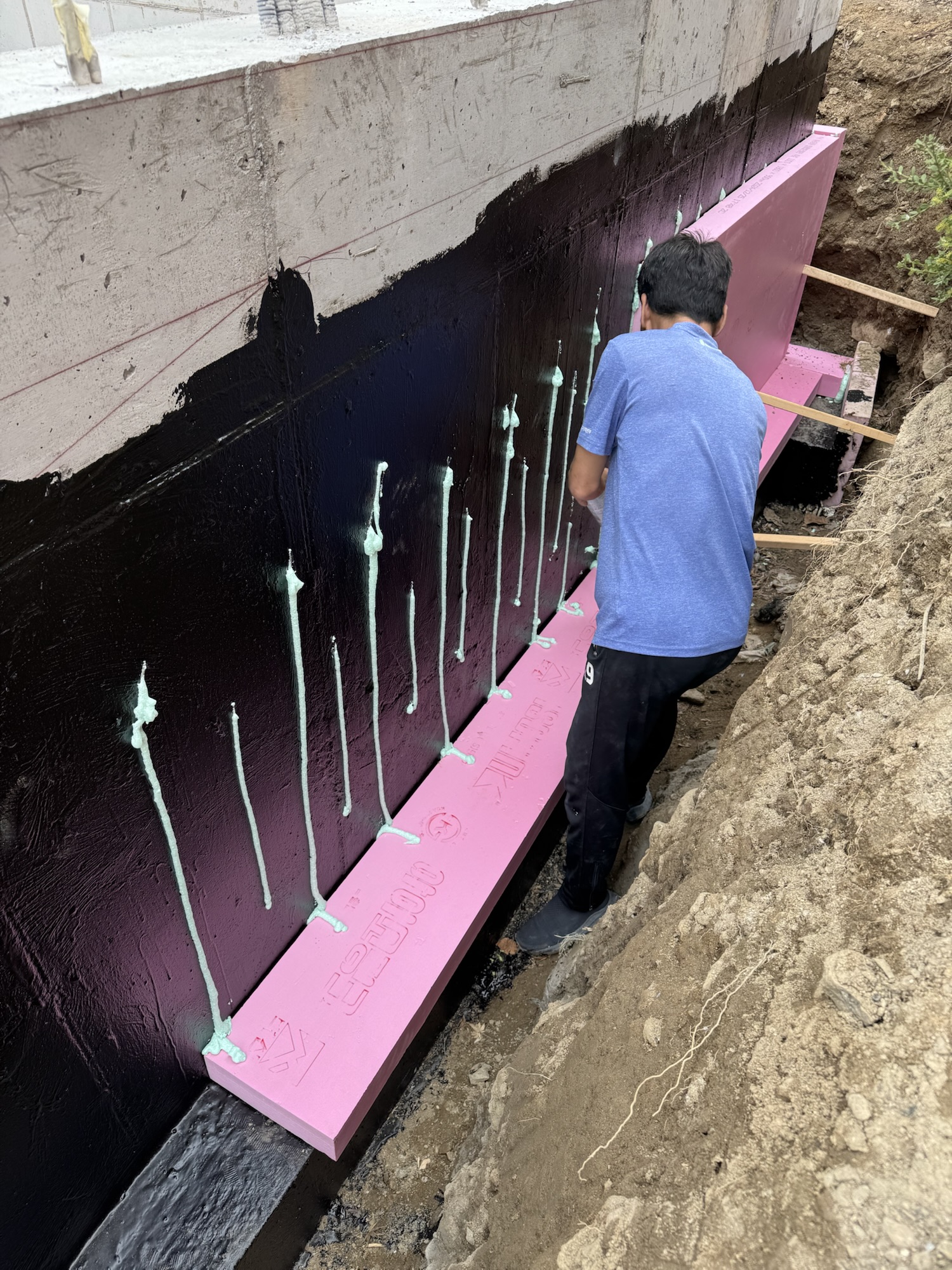 공사 현장에서 방수 처리를 위해 벽체에 방수제를 바르고 있는 작업자. 벽체 옆에는 단열재가 부착되고 있으며, 주위에는 흙이 둥글게 쌓여 있다.