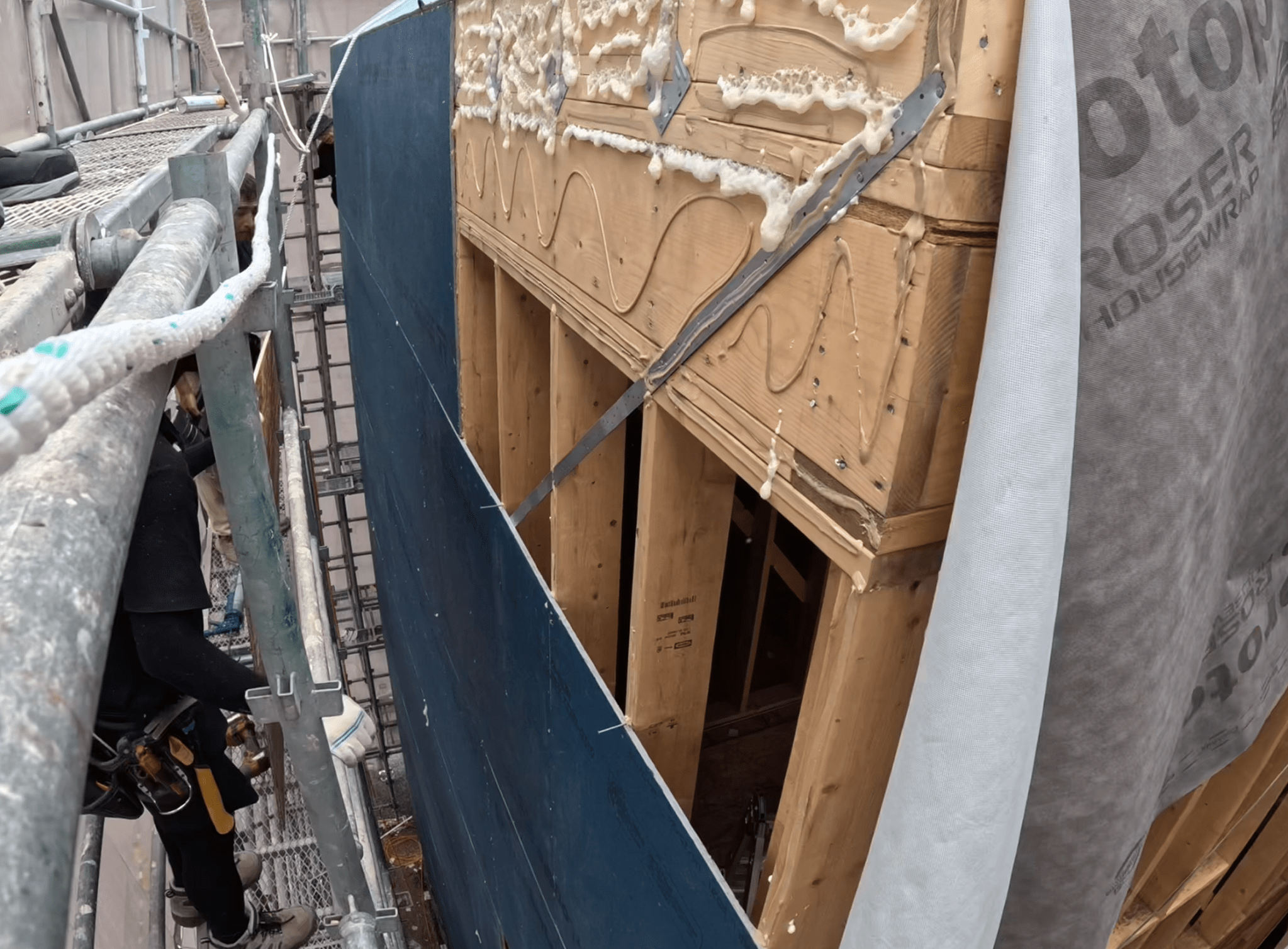 건축 현장에서 접착제를 사용하여 벽체 시공 중인 모습, 스캐폴딩이 보이고, 나무 프레임과 접합부에 접착제가 발라져 있음.