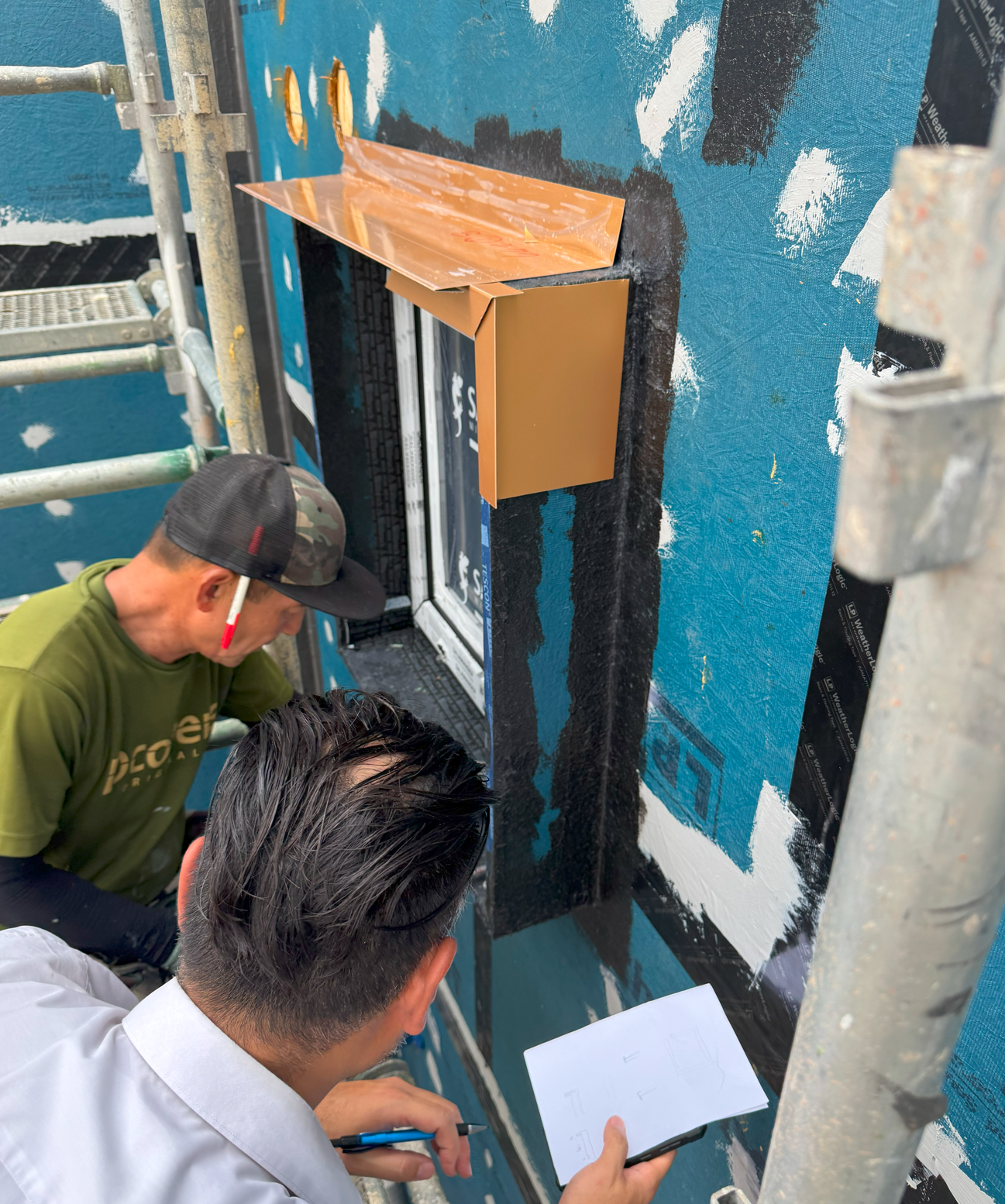 두 명의 남성이 창문 주변에서 작업하며, 메모와 자료를 참고하고 있는 모습. 외부 벽면은 파란색으로 칠해져 있으며, 창문 위에는 금속 플래싱이 설치되고 있다.