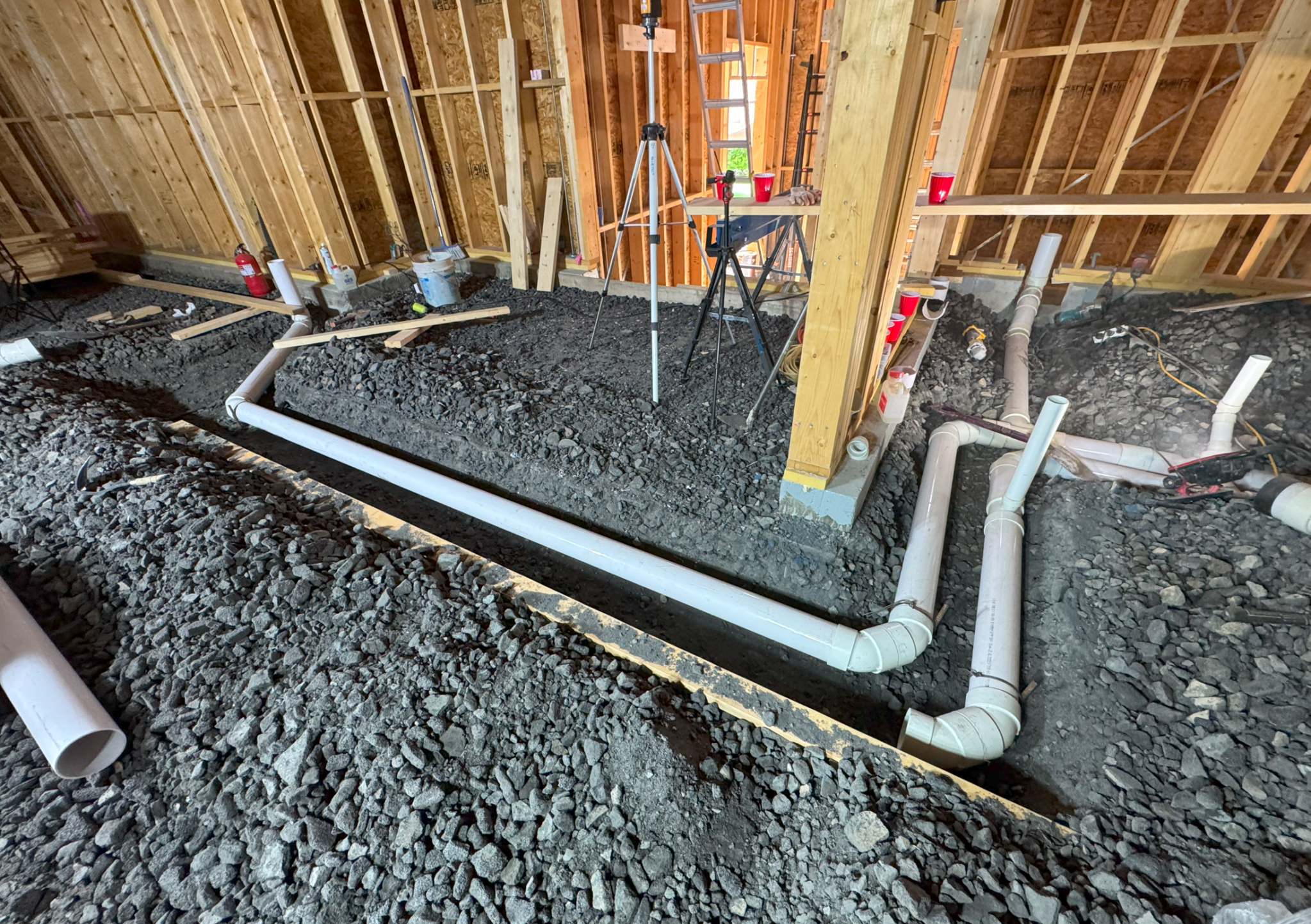 건축 현장의 내부 모습으로, 바닥에 자갈과 배수 및 하수관이 설치되어 있는 장면을 보여줍니다. 벽에는 나무 프레임이 보이며, 공구와 장비들이 배치되어 있습니다.