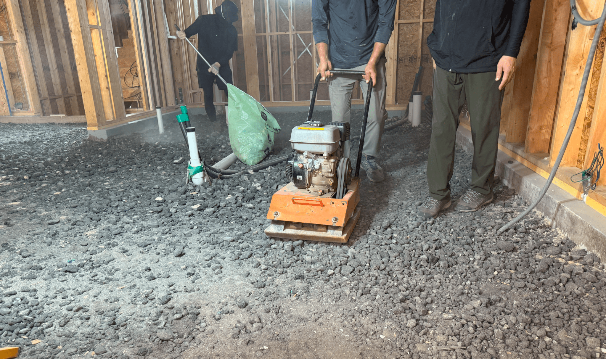 Construction workers compacting gravel with a plate compactor in an unfinished indoor area.
