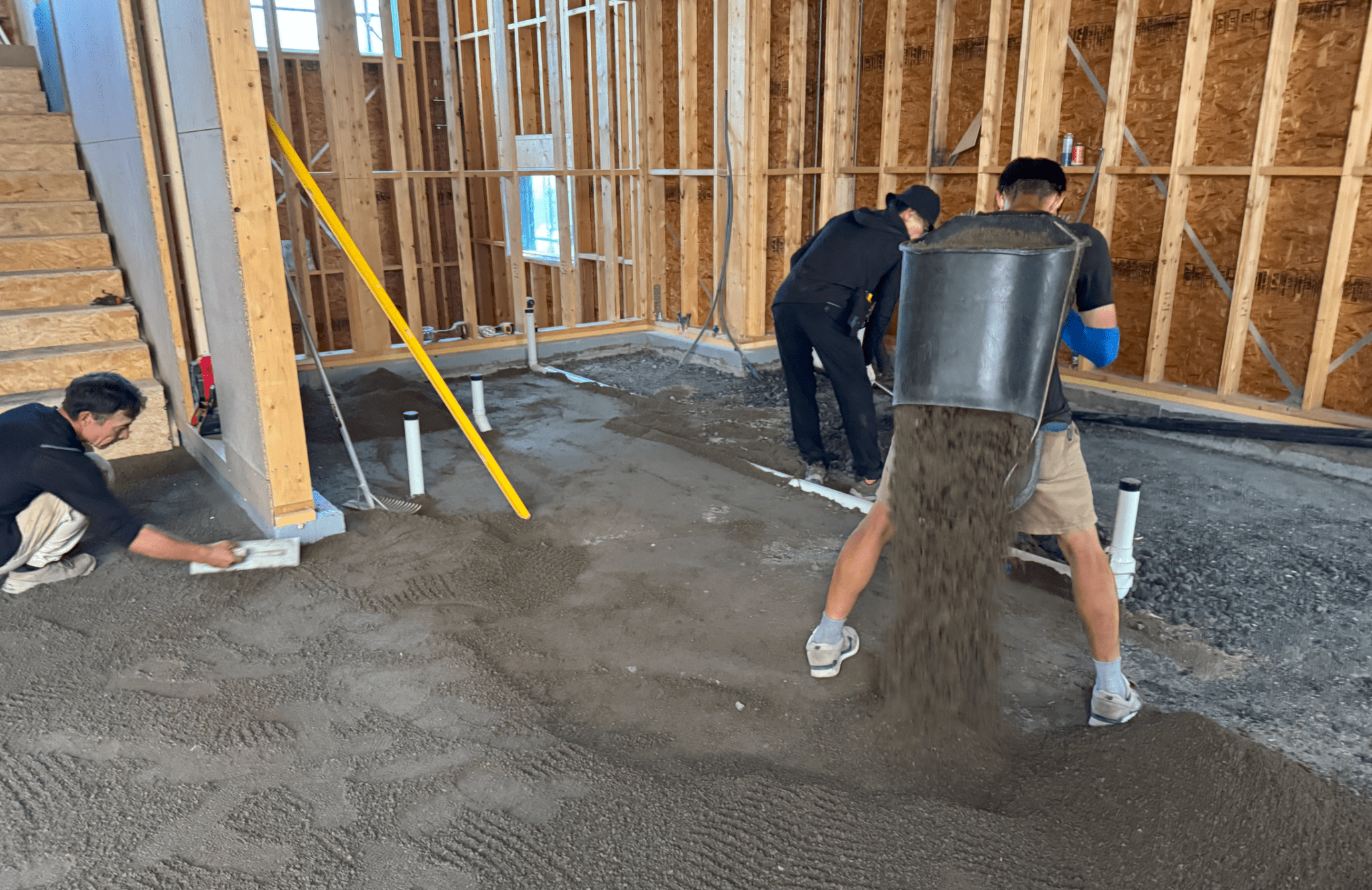 Construction workers leveling a floor with aggregate in a building under construction, featuring wooden framing and plumbing pipes.