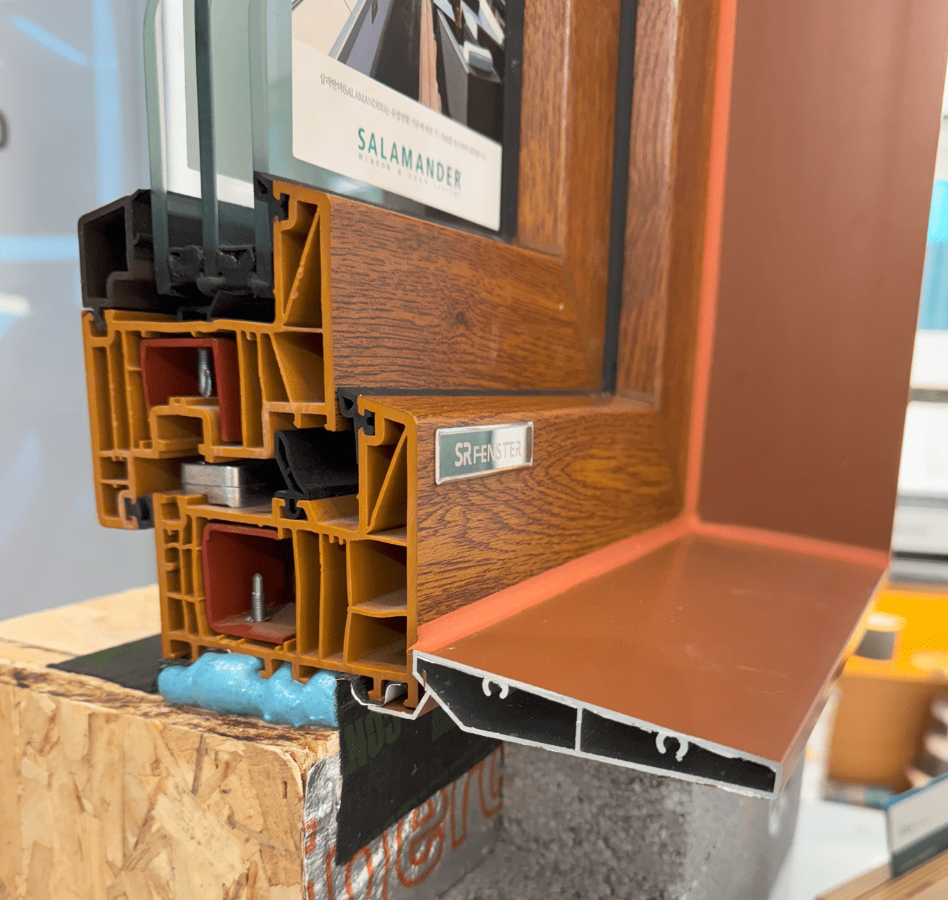 SRFENSTER 브랜드의 다층 구조 유리창호 조각, 나무 패턴의 외관과 알루미늄 플래싱이 결합된 디자인.