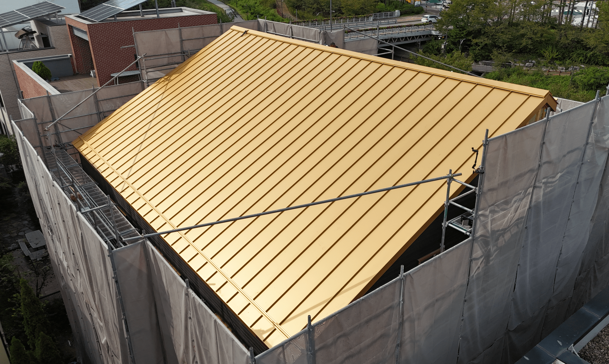 금색의 스탠딩 심 지붕이 있는 건물의 위쪽 전망. 지붕은 기둥과 비계에 의해 둘러싸여 있고, 주변에 나무와 건축물 일부가 보인다.
