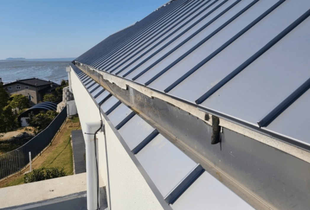 A close-up view of a metal roof edge showing a gutter system and a coastal landscape in the background.