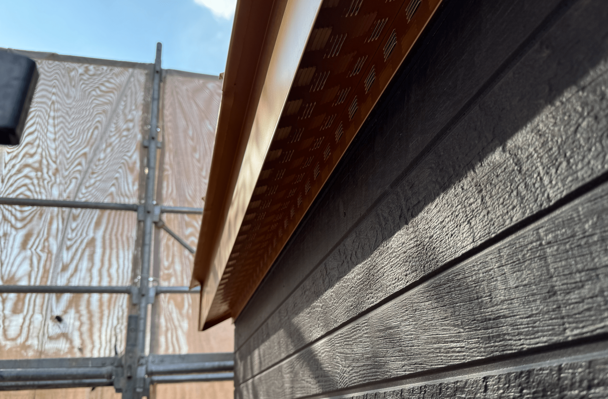 지붕의 가장자리가 보이는 이미지로, 금속 재질의 거터 시스템과 검은색 외벽이 함께 나타나 있으며, 청명한 하늘이 배경에 있습니다.